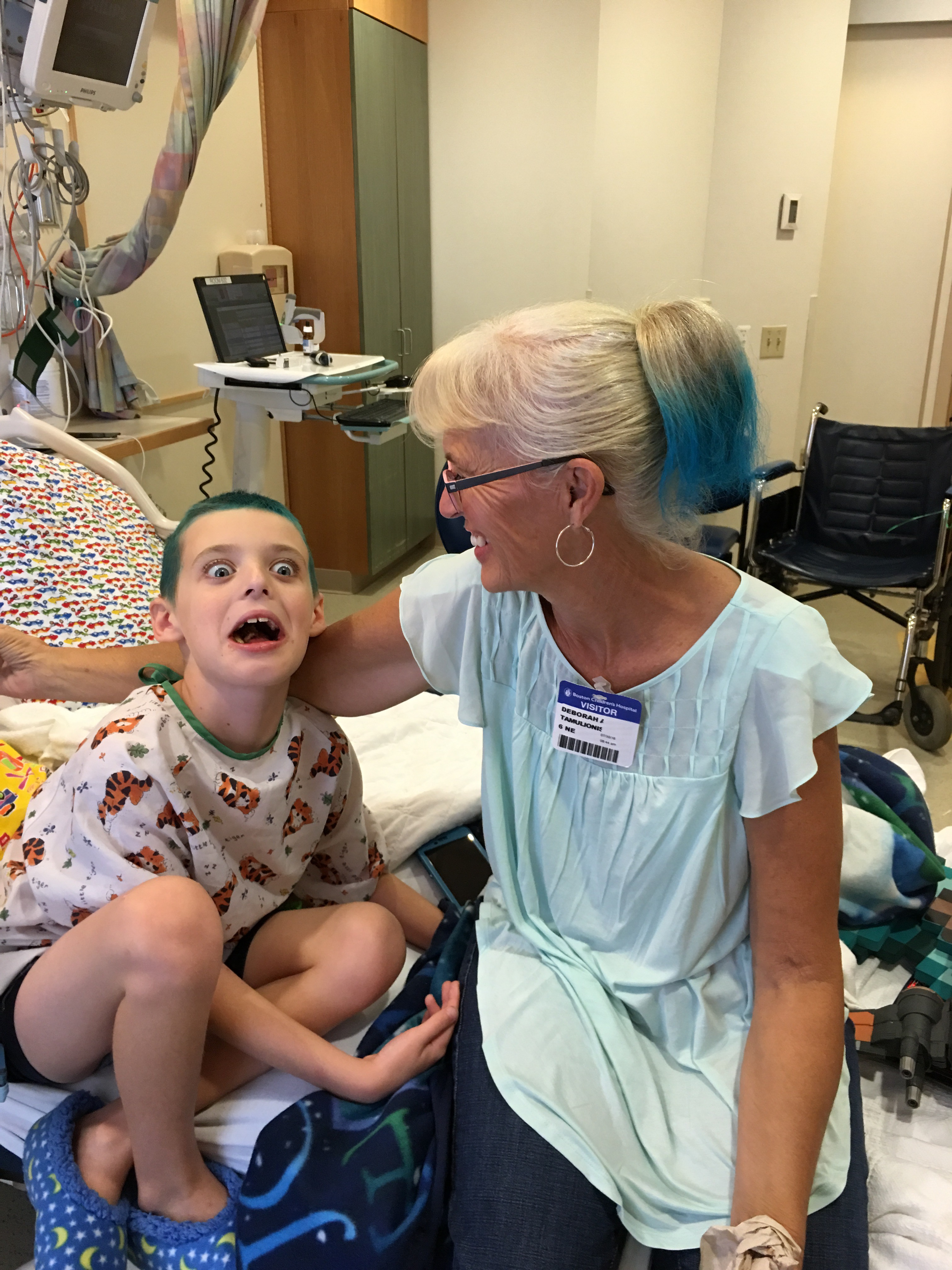 Max sitting up and smiling wide in a hospital bed after dying his hair green